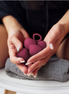 A person holds a small cluster of matte pink silicone personal care items in both hands over a gray towel, with a black top visible in the background.